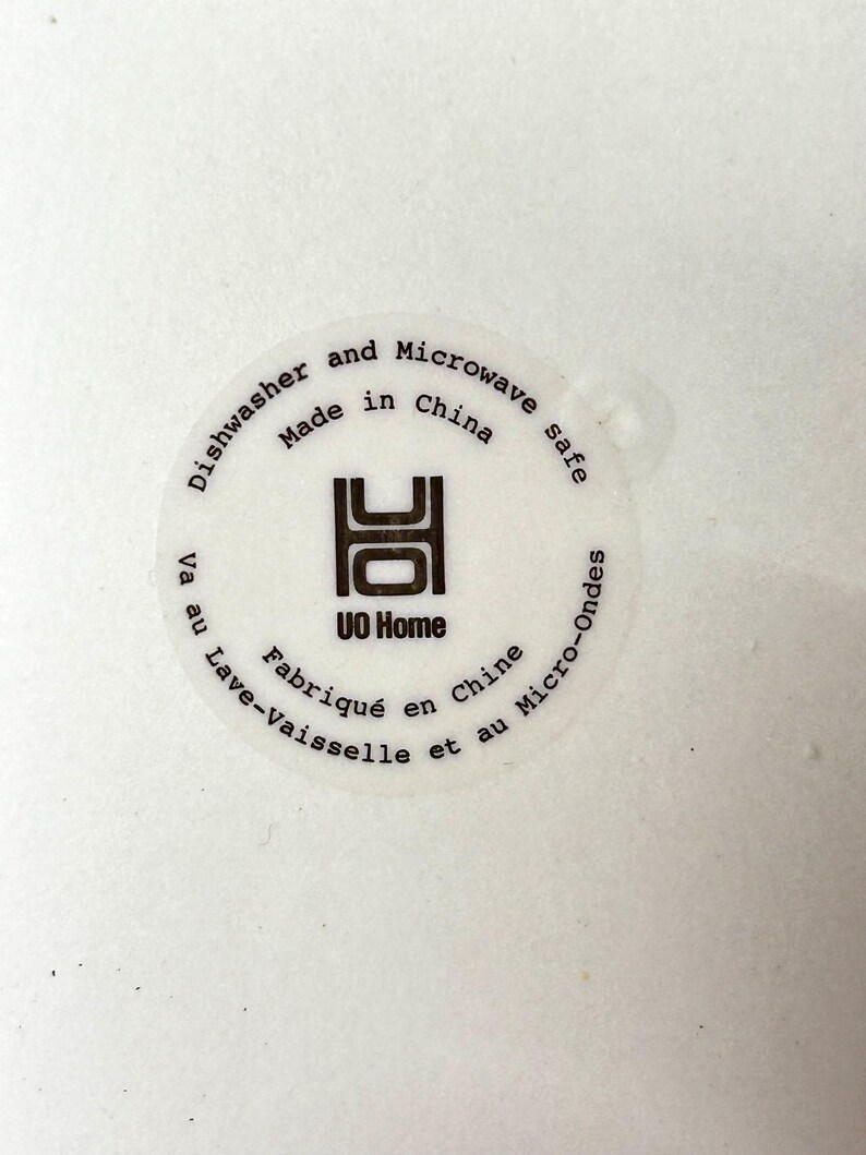 Puede incluir: Primer plano de una superficie blanca con una pegatina circular. La pegatina dice "Dishwasher and Microwave safe Made in China" y "UO Home". Tambi&eacute;n hay texto en franc&eacute;s: "Fabriqu&eacute; en Chine" y "Lave-Vaisselle et au Micro-Ondes".