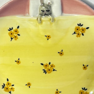 Puede incluir: Plato de cer&aacute;mica con dise&ntilde;o de conejito durmiendo. El plato presenta una manta amarilla con estampados florales amarillos, una cabecera rosa y una almohada blanca. El conejito es gris y parece estar durmiendo.
