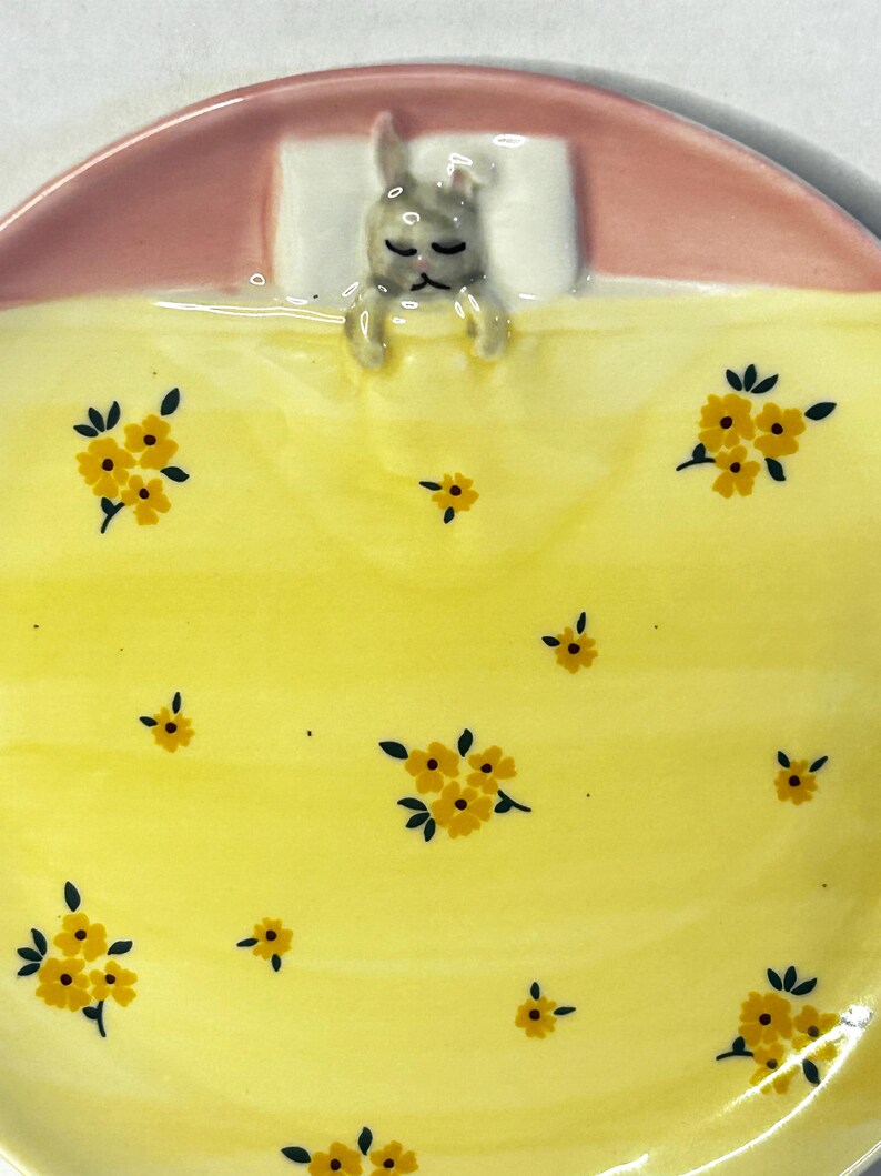 Puede incluir: Plato de cer&aacute;mica con dise&ntilde;o de conejito durmiendo. El plato presenta una manta amarilla con estampados florales amarillos, una cabecera rosa y una almohada blanca. El conejito es gris y parece estar durmiendo.