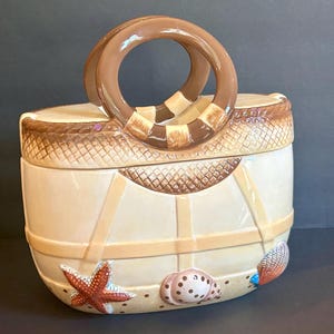 May include: A ceramic cookie jar shaped like a beach tote bag with a cream-colored body, brown circular handles, and a woven texture pattern at the top. The base is decorated with a raised red starfish, a spotted seashell, and a blue-tinted shell.