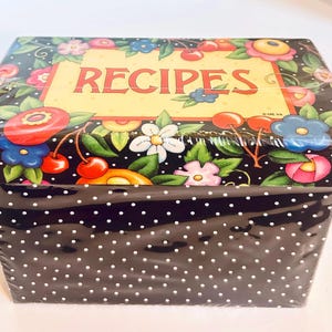 May include: A rectangular recipe box with a black base featuring white polka dots. The lid has a yellow center panel with the word RECIPES in red letters, surrounded by a colorful border of red cherries, green leaves, and various flowers.