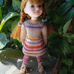 Op de afbeelding: Een pop met rood haar in vlechten, gekleed in een gebreide jurk met een kleurrijke gestreepte rok en een patroon lijfje. De pop draagt ook roze leggings en bruine laarzen. De achtergrond toont groen gebladerte en een stenen oppervlak.