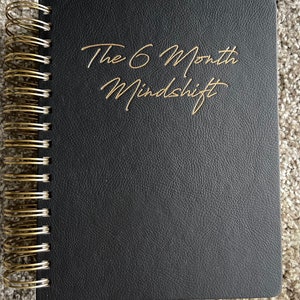 May include: A black leather-bound journal with a gold spiral binding. The cover has the text "The 6 Month Mindshift" written in gold lettering.