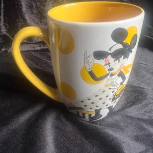 Puede incluir: Taza de cerámica blanca con interior y asa amarillos. La taza presenta un personaje de dibujos animados con gafas de sol y una bufanda amarilla, con lunares amarillos en el exterior. Un artículo divertido y elegante.
