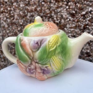 Pode incluir: Uma chaleira de cerâmica com tampa, decorada com um desenho de vegetais coloridos. A chaleira é branca com vegetais verdes, amarelos, laranja e roxos.