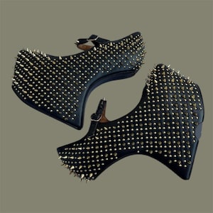 May include: Black platform shoes covered in gold-coloured spikes. The shoes feature a Mary Jane design with a strap and buckle. The surface of the shoes is densely covered in small, pointed spikes.
