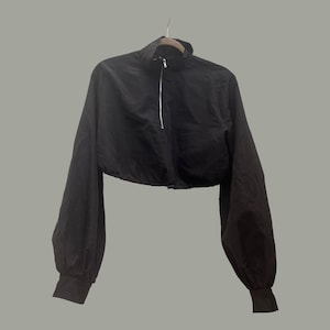 May include: A black cropped jacket with a high collar and long sleeves. The jacket has a silver zipper down the front and is hanging on a wooden hanger. The sleeves have a gathered, balloon-like shape and end in fitted cuffs.