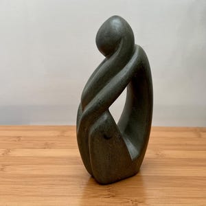 May include: A dark green sculpture of a person in a seated, curled position. The figure's arms are wrapped around the knees, and the head is bowed. The sculpture is on a wooden surface.