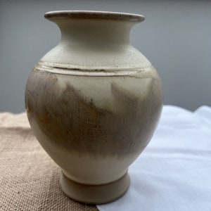May include: A small, cream-colored ceramic vase with a brown glaze. The vase has a rounded body and a flared lip.
