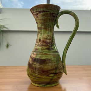 Op de afbeelding: Een groene en bruine keramische vaas met een brede opening en een gebogen handvat. De vaas heeft een swirl-patroon.