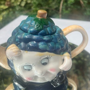 Puede incluir: Una tetera de cerámica azul y blanca con forma de oveja con una hoja verde en la parte superior. La tetera tiene una cara caprichosa con ojos grandes y una sonrisa.