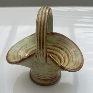 May include: A small, green ceramic basket with a brown handle. The basket has a swirl pattern and is handmade.