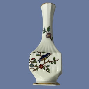 Op de afbeelding: Een witte keramische vaas met een vogel- en bloemenontwerp. De vaas heeft een slanke hals en een bredere, facetgeslepen body. De vogel is blauw, geel en bruin, op een tak met rode en blauwe bloemen en groene bladeren. Gouden afwerking.