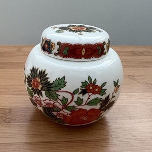 May include: White ceramic jar with a lid featuring a floral design in shades of red, green, and black. The jar has a round shape and a wide mouth.