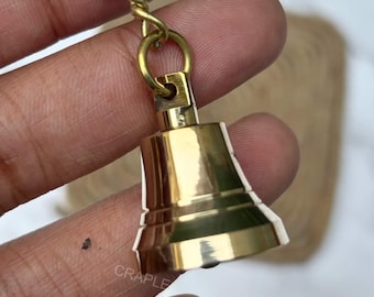 May include: A small, shiny brass bell keychain. The bell is a classic shape with a loop at the top connected to a chain and keyring. The bell is held in a hand against a blurred background.