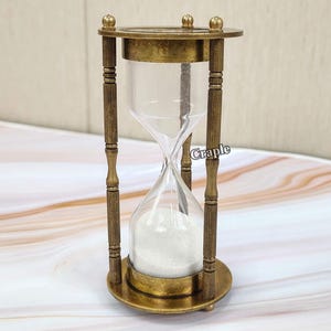 以下が含まれることがあります： 真鍮フレームと透明なガラスのアンティークスタイルの砂時計。砂時計には白い砂が入っており、4本の木の柱で支えられています。上部と下部は円形で、装飾的なディテールがあります。「Craple」という単語が見えます。