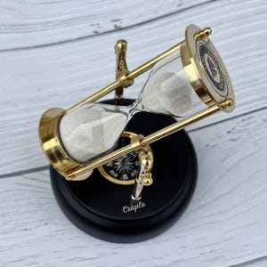 Può includere: Una clessidra con cornice dorata e base nera. La camera di vetro contiene sabbia bianca. La base ha la scritta "Craple". La clessidra è inclinata su una superficie di legno chiaro.