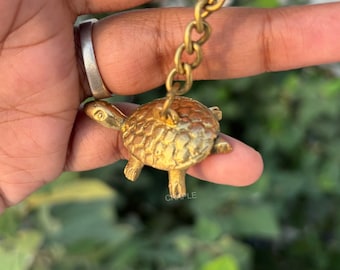 May include: A small, detailed brass turtle keychain. The turtle has a textured shell and is attached to a chain and keyring. The keychain is held against a blurred green background.