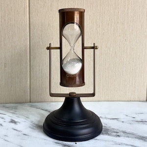 May include: A vintage hourglass with a bronze-coloured frame and a black base. The glass chamber contains white sand, and the word "Craple" is visible on the glass. The hourglass is supported by a metal frame.