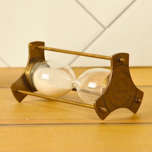 May include: A brass hourglass with white sand, featuring a circular base with the word "Crooked" inscribed on it.