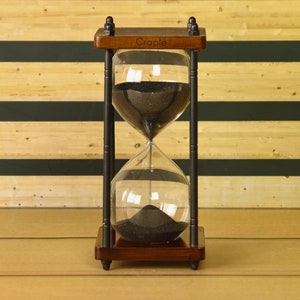 May include: A wooden hourglass with black sand and a black metal frame. The hourglass is on a wooden surface and has the word "Craple" engraved on the top.