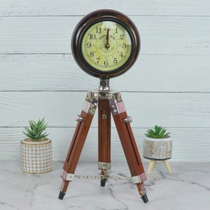 May include: A vintage-style clock with a cream-colored face and black numerals sits atop a wooden tripod stand. The clock has a dark brown bezel and silver-colored metal accents. The tripod legs are dark brown with pink accents.