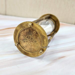 May include: An antique brass hourglass with a compass rose design on the base. The hourglass has a clear glass center and is filled with white sand. The compass rose features directional markings.