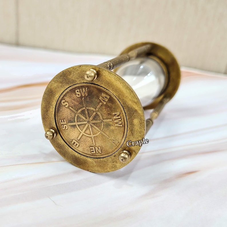 May include: An antique brass hourglass with a compass rose design on the base. The hourglass has a clear glass center and is filled with white sand. The compass rose features directional markings.