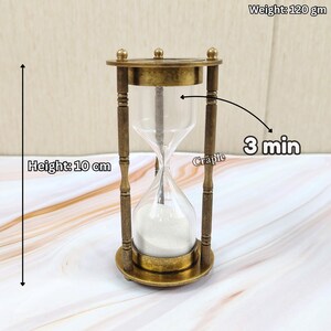 May include: A vintage-style hourglass with a brass frame and clear glass. The hourglass is 10 cm tall and weighs 120 grams. The sand timer is designed to measure 3 minutes. The hourglass has a decorative design.