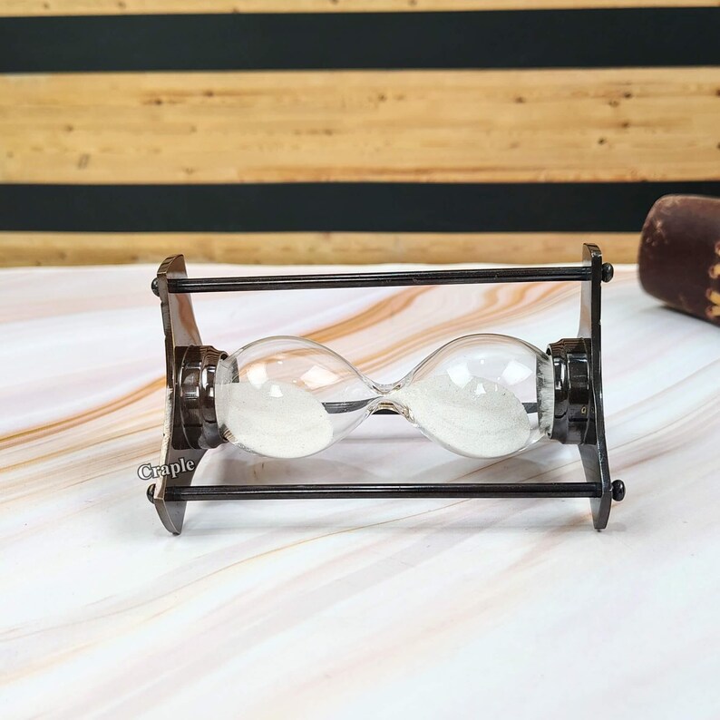 May include: An hourglass with clear glass bulbs and white sand, set in a dark brown metal frame. The hourglass is on a white and gray marble-patterned surface. The word "Craple" is printed on the frame.