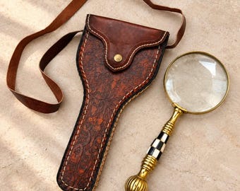 May include: A brown leather case with an embossed floral pattern and a long strap, paired with a magnifying glass featuring a gold-toned handle with a black and white checkered pattern and a round glass lens set in a gold-toned metal frame.