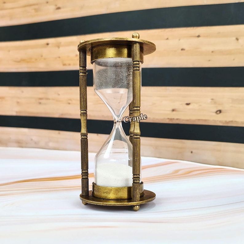 May include: An antique-style hourglass with a brass-coloured metal frame and clear glass chambers. The hourglass is filled with white sand. The design includes decorative vertical bars and a circular base. The background features a wooden panel.