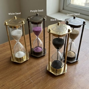 Könnte beinhalten: Vier sechseckige Sanduhren mit Metallrahmen und Glaskammern. Jede enthält unterschiedlich farbigen Sand, beschriftet mit White Sand, Purple Sand, Black Sand und Desert Sand. Die Deckplatten zeigen ein geprägtes Schiffsmotiv und den Text The Mary Rose 1511-1545.