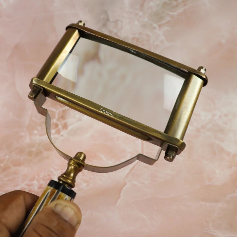May include: A vintage brass magnifying glass with a rectangular lens. The handle features a decorative design with a patterned grip. The frame is a warm, antique gold color, and the lens is clear.