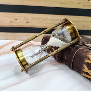 Op de afbeelding: Een antieke messing zandloper met heldere glazen kamers gevuld met wit zand. De zandloper heeft een klassiek ontwerp met decoratieve messing steunen en eindkappen. Het woord "Craple" is op het glas gedrukt. De zandloper staat tegen een bruine lederen achtergrond.