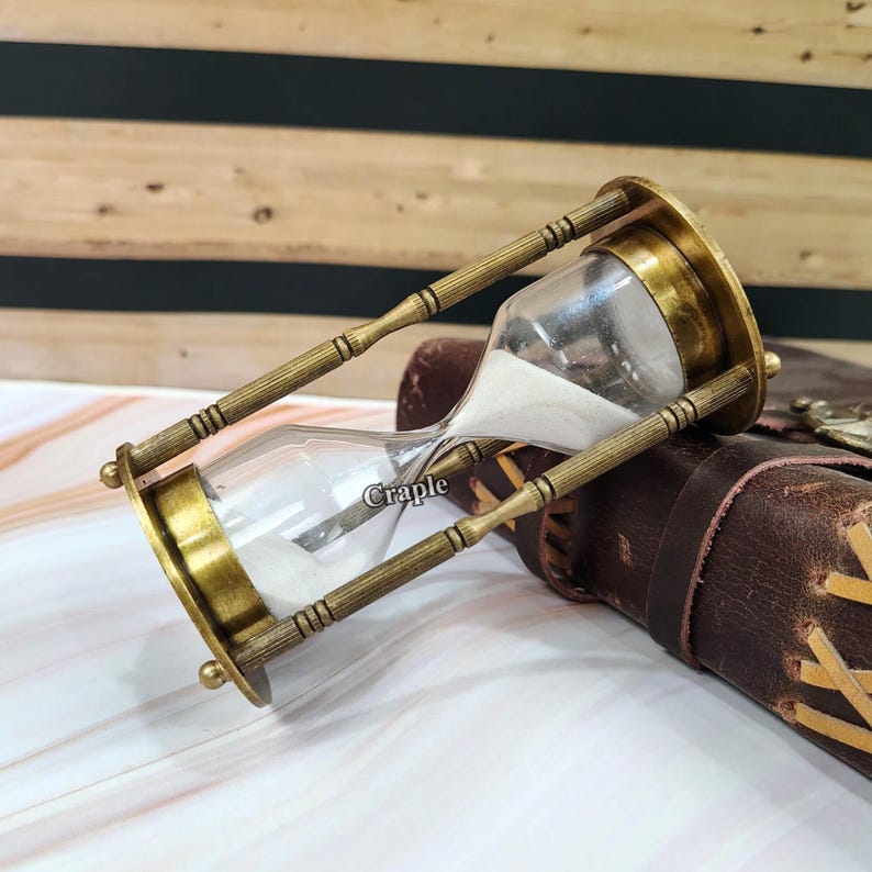 May include: An antique brass hourglass with clear glass chambers filled with white sand. The hourglass has a classic design with decorative brass supports and end caps. The word "Craple" is printed on the glass. The hourglass is set against a brown leather background.
