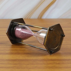 May include: A brass hourglass with a hexagonal base and a clear glass bulb filled with pink sand. The hourglass is decorated with an inscription that reads "1851 The First Ship To Cross The Atlantic"