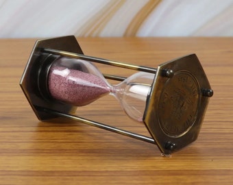 May include: A brass hourglass with a hexagonal base and a clear glass bulb filled with pink sand. The hourglass is decorated with an inscription that reads "1851 The First Ship To Cross The Atlantic"