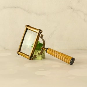 May include: A vintage-style brass magnifying glass with a rectangular lens. The handle has a ribbed design and a black end. The word "Craple" is printed on the frame. The magnifying glass is resting on a small green and white pot.