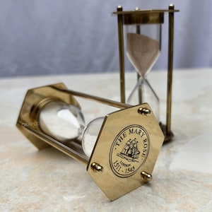 May include: Two brass hourglasses with clear glass chambers. One is lying on its side, displaying the engraved text "THE MARY ROSE LONDON 1511-1545". The other stands upright, with sand flowing through it.