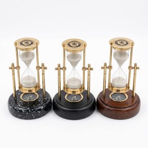 May include: Three hourglasses with brass frames and white sand, each featuring a compass embedded in the base and a top plate inscribed with Ross London 1917. The bases are finished in speckled black, matte black, and polished dark brown wood.