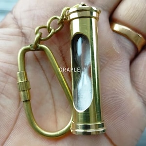 May include: Brass hourglass keychain with a white sand timer inside. The keychain has a brass loop and a brass chain.