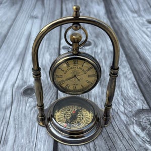 May include: Antique brass pocket watch and compass set. The watch face displays Roman numerals and the words "Victoria London 1876". The compass is visible below the watch. The set is mounted on a decorative stand.