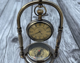 May include: Antique brass pocket watch and compass set. The watch face displays Roman numerals and the words "Victoria London 1876". The compass is visible below the watch. The set is mounted on a decorative stand.