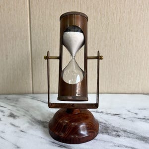 Può includere: Una clessidra vintage con una cornice in metallo color rame e una base in legno. La camera di vetro contiene sabbia bianca. La clessidra è sostenuta da una cornice in metallo con un design curvo. La parola "Crape" è stampata sul vetro.