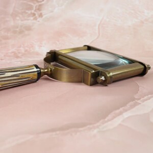 May include: A vintage-style brass magnifying glass with a rectangular lens. The handle features a cream and brown striped pattern. The magnifying glass has a curved handle and a brass frame. The word "Craple" is printed on the side of the frame.