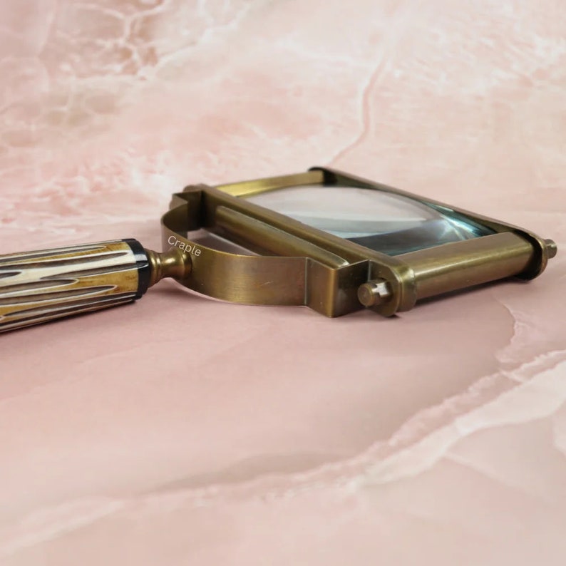 May include: A vintage-style brass magnifying glass with a rectangular lens. The handle features a cream and brown striped pattern. The magnifying glass has a curved handle and a brass frame. The word "Craple" is printed on the side of the frame.