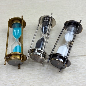 May include: Three hourglass sand timers with different colored sand. The hourglasses are made of brass and have a silver finish. The sand is blue, black, and white.