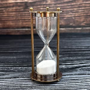 May include: A brass hourglass with a clear glass bulb and white sand. The hourglass is on a dark wooden surface. The base of the hourglass has the word "CRAPLE" inscribed on it.