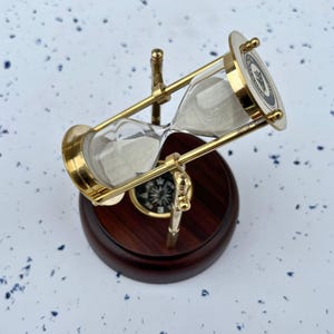 May include: An hourglass with a brass frame and a wooden base. The glass contains white sand. A compass is visible on the wooden base. The hourglass is tilted, showing the sand flowing from the top to the bottom.